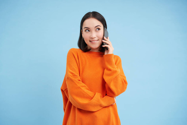 Корейская девушка в оранжевом свитере разговаривает по мобильному телефону, говорит по телефону с счастливым улыбающимся лицом, заказывает доставку, консультируется с кем-то по телефону. - Фото, изображение