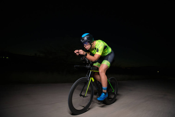 A triathlete rides his bike in the darkness of night, pushing himself to prepare for a marathon. The contrast between the darkness and the light of his bike creates a sense of drama and highlights the - Foto, Bild