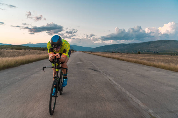  Triathlete riding his bicycle during sunset, preparing for a marathon. The warm colors of the sky provide a beautiful backdrop for his determined and focused effort - Photo, Image
