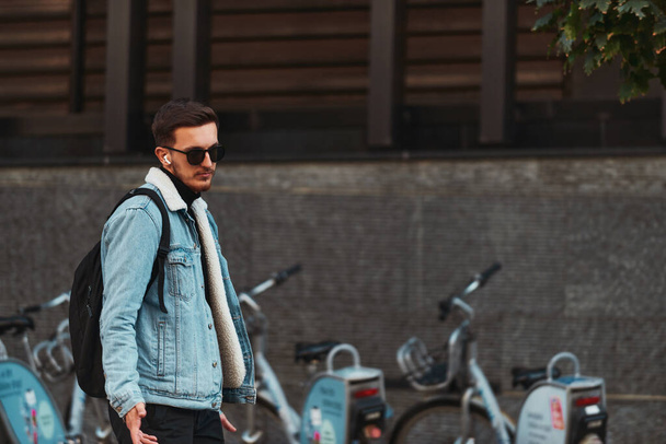Студент прогуливается по городу в солнечных очках, носит рюкзак и использует беспроводные наушники, воплощая современный городской образ жизни и возможность оставаться на связи во время поездки - Фото, изображение