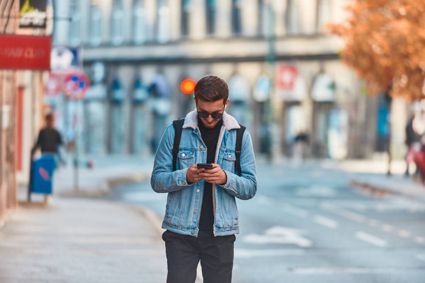 Студент, гуляющий по городу с рюкзаком и в солнечных очках во время использования смартфона, представляющий современный городской образ жизни и полагающийся на технологии для коммуникации и - Фото, изображение