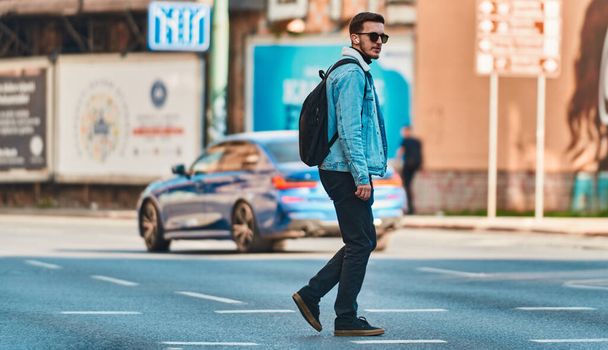 Студент прогуливается по городу в солнечных очках, носит рюкзак и использует беспроводные наушники, воплощая современный городской образ жизни и возможность оставаться на связи во время поездки - Фото, изображение