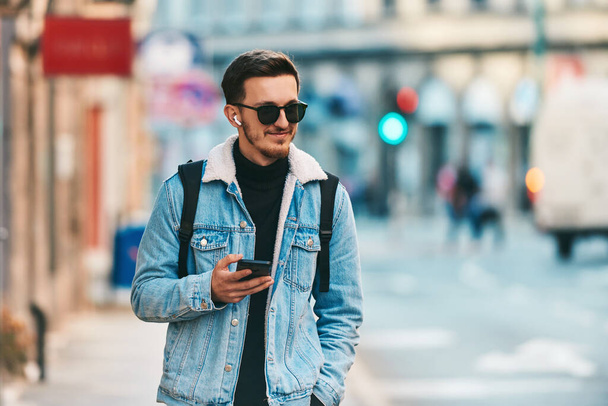 Un étudiant se promène dans la ville en portant un sac à dos et des lunettes de soleil tout en utilisant un smartphone, représentant le mode de vie urbain moderne et la dépendance à la technologie pour la communication et - Photo, image