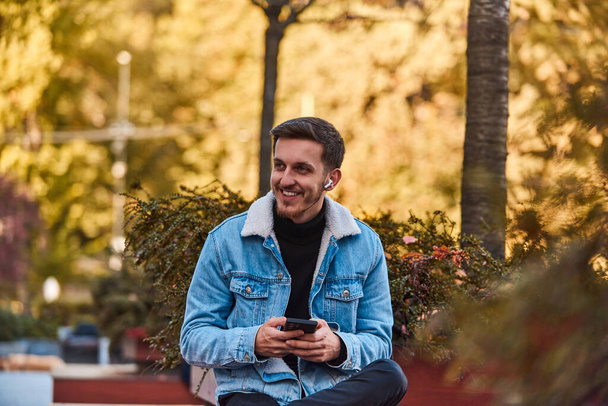 A man sitting on a bench in the city center using his smartphone and wireless headphones for a call, showcasing the convenience of technology in modern communication and the ability to multitask while - Photo, Image