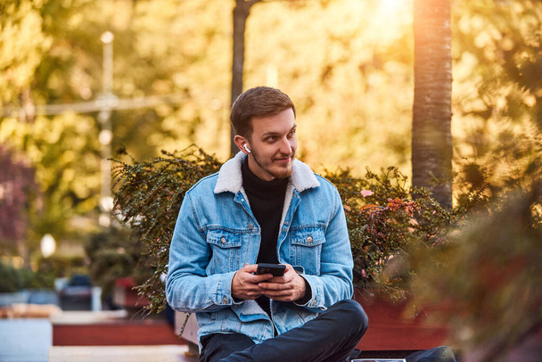 Mies istuu penkillä kaupungin keskustassa käyttäen älypuhelinta ja langattomia kuulokkeita puheluun, jossa esitellään teknologian mukavuutta nykyaikaisessa viestinnässä ja kykyä monitehtävään - Valokuva, kuva