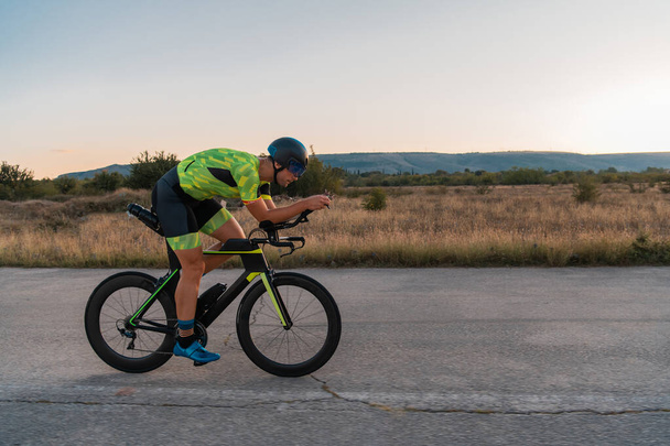  Triathlete riding his bicycle during sunset, preparing for a marathon. The warm colors of the sky provide a beautiful backdrop for his determined and focused effort - Foto, afbeelding