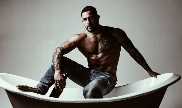 That means its bath time. Strong latino man with tattoo on muscular torso sitting in bath. Atheltic hispanic man drinking wine in bath. Wine and bath are the key to relaxation for him. - Photo, Image