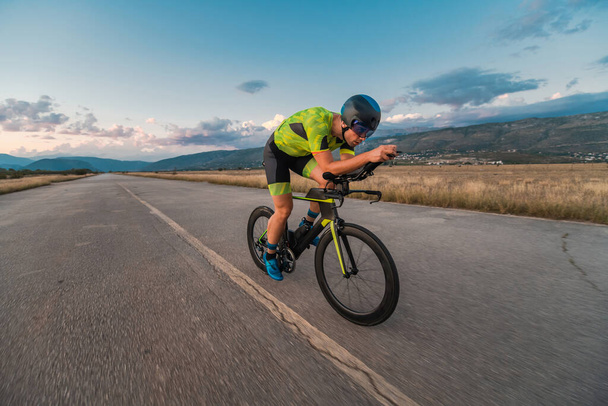  Triathlete riding his bicycle during sunset, preparing for a marathon. The warm colors of the sky provide a beautiful backdrop for his determined and focused effort - Φωτογραφία, εικόνα