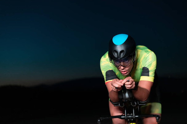 A triathlete rides his bike in the darkness of night, pushing himself to prepare for a marathon. The contrast between the darkness and the light of his bike creates a sense of drama and highlights the - Zdjęcie, obraz