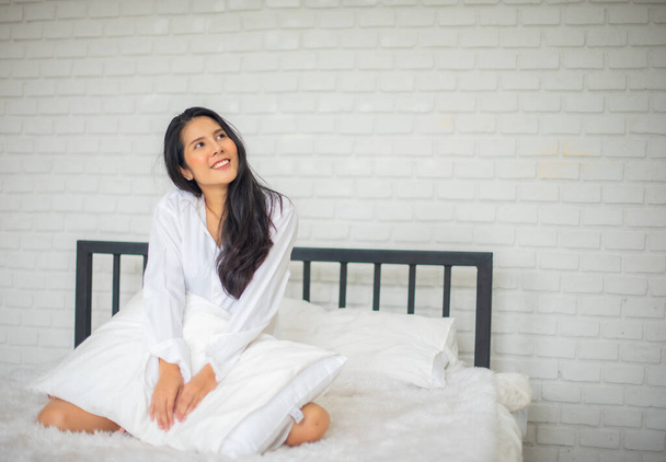 Černovlasá žena sedící ráno v luxusním pokoji na posteli s oběma rukama v podušce a usmívá se na kopírovací prostor s úsměvem na tváři. - Fotografie, Obrázek
