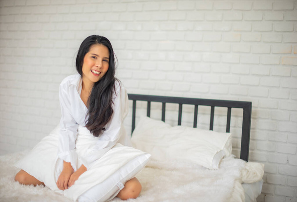 Black haired woman sitting of a modern hotel room in the morning on the bed with both hands holding the pillow and smile at the camera. - Photo, Image