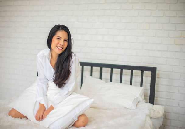 Black haired woman sitting of a luxury modern hotel room in the morning on the bed with both hands holding the pillow and smiles at the camera. - Fotoğraf, Görsel