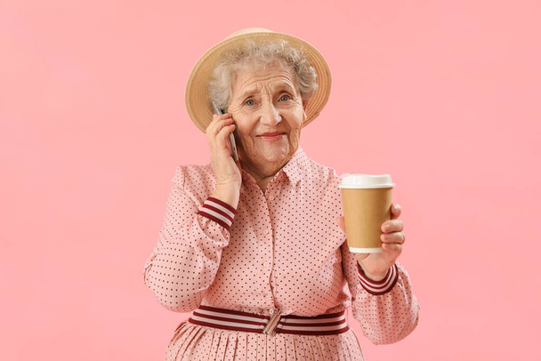 Senior woman with cup of coffee talking by mobile phone on pink background - Photo, Image