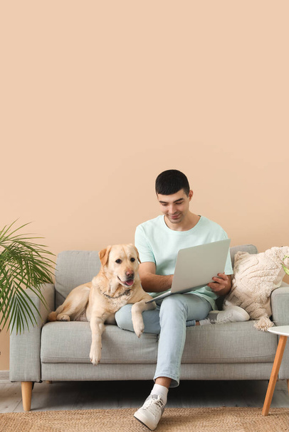 Νεαρός με φορητό υπολογιστή και το Λαμπραντόρ σκυλί του κάθεται στον καναπέ στο σπίτι - Φωτογραφία, εικόνα