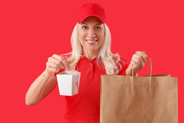 Ώριμος κούριερ με κουτί τροφίμων και χάρτινη σακούλα σε κόκκινο φόντο - Φωτογραφία, εικόνα