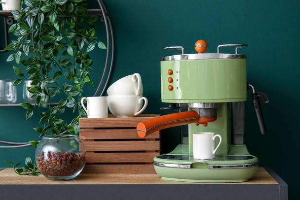 Современная кофеварка, чашки и банка с бобами на столе у зеленой стены - Фото, изображение