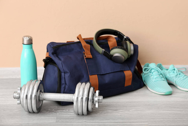Спортивная сумка с наушниками, кроссовками, гантели и бутылка возле бежевой стены в тренажерном зале - Фото, изображение