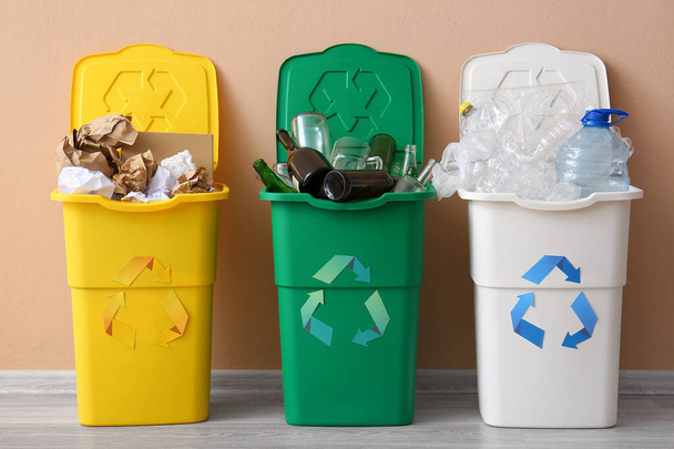 Сміттєві баки з символом переробки та різними сміттями біля бежевої стіни
 - Фото, зображення