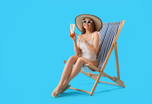 Belle jeune femme avec crème solaire assise sur une chaise longue sur fond bleu - Photo, image