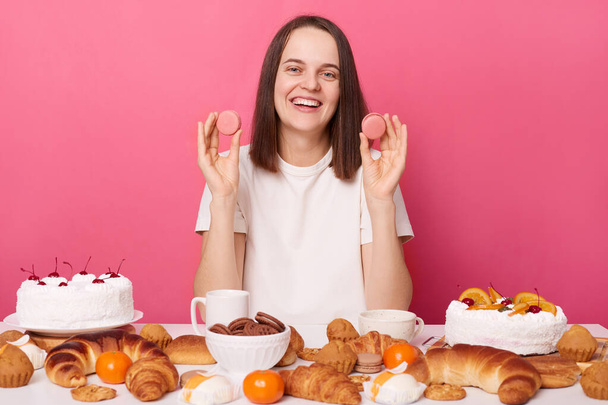 Smiling pleased pretty brown haired woman in white t shirt sitting at table with desserts isolated over pink background holding cookies enjoying sweet dinner. - Foto, Imagen