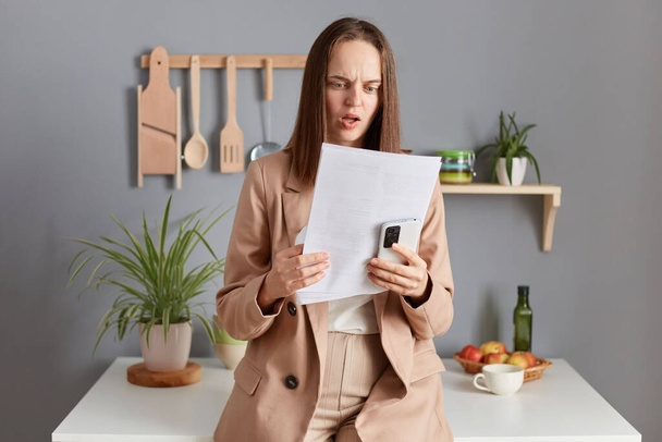 Shocked surprised disappointed woman with brown hair wearing beige jacket standing in home kitchen interior standing with smart phone in hands reading papers with astonishment. - 写真・画像