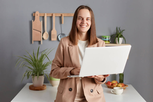 Pleased smiling woman freelancer with brown hair wearing beige jacket standing in kitchen interior working on laptop from home preparing for online meeting. - Photo, image