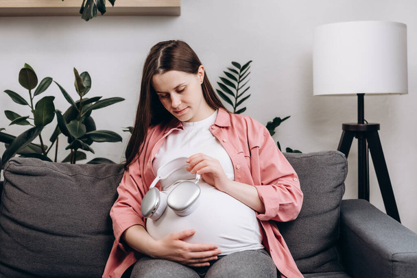 Loving happy expectant mother listening to music song melody enjoying pregnancy sitting on cozy sofa at home, taking off headphones from head putting on pregnant belly audio therapy unborn baby - Valokuva, kuva