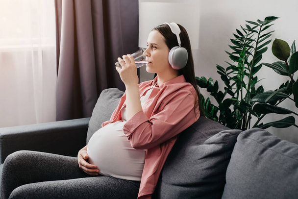 Relaxed beautiful young pregnant female in wireless headphones listening to music sitting on comfy sofa at home and drinking glass of fresh water in living room. Pregnancy and melody therapy concept - Valokuva, kuva