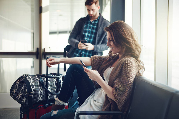 Απλά αποχαιρετώ την οικογένεια. ένα νεαρό ζευγάρι που κάθεται σε ένα αεροδρόμιο με τις αποσκευές τους ενώ χρησιμοποιούν τα κινητά τους τηλέφωνα - Φωτογραφία, εικόνα