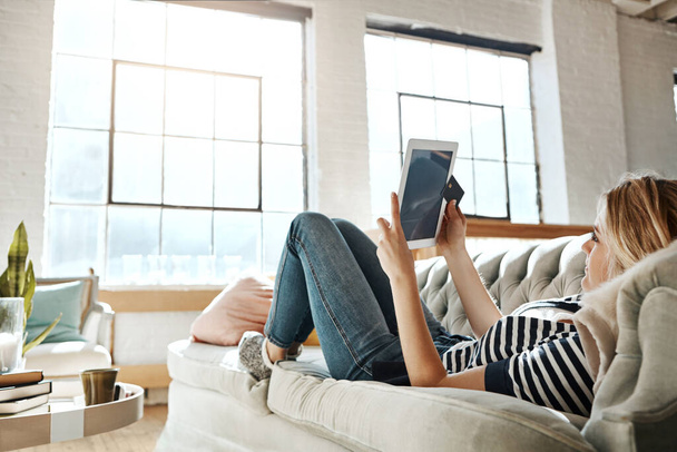 Kytketty virtuaalikauppaan. Nuori nainen rentoutuu sohvalla ja käyttää luottokorttia digitaalisen tabletin kanssa - Valokuva, kuva