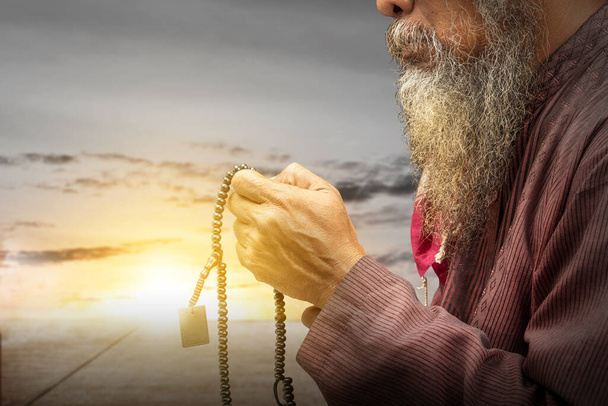 Muslim man with a beard wearing keffiyeh with agal praying with prayer beads on his hands with the sunset scene background - Fotó, kép