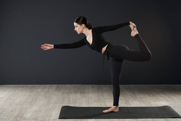 Zijaanzicht van een jonge vrouw die blootsvoets op een yoga mat staat en traint. Slanke, brunette vrouw met sportkleding, stretching, benen optillen. Geïsoleerd op zwarte studio achtergrond. - Foto, afbeelding