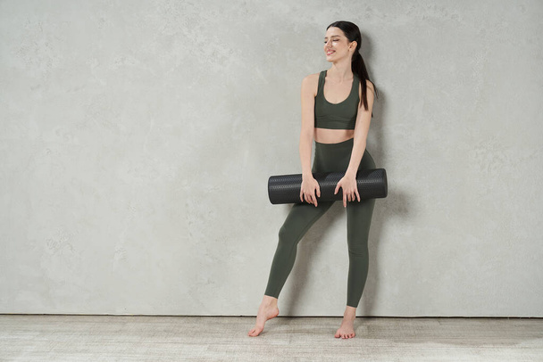Front view of barefoot woman wearing sportswear, working out, having break. Young, slim female standing, looking aside, smiling, holding yoga mat. Concept of exercising and training. - Fotografie, Obrázek