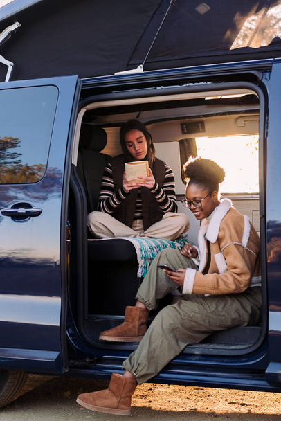 two young women enjoying camper van life reading at sunset, concept of travel adventure and female friendship - Foto, Bild