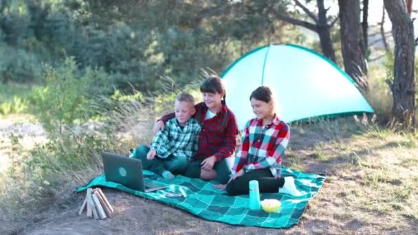 Bambini in cerca di spettacolo sul computer portatile nella foresta. Ragazzi e ragazze stanno riposando in un campo tenda. I bambini si siedono sul plaid sull'erba in estate. - Filmati, video