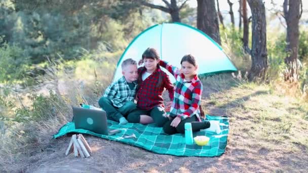 Children looking show on laptop in the forest. Boys and girls are resting in a tent camp. Children sit on the plaid on the grass in summer. - Footage, Video