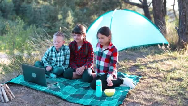Enfants regardant spectacle sur ordinateur portable et manger du maïs pop dans la forêt. Garçons et filles se reposent dans un camp de tentes. Les enfants s'assoient sur le plaid sur l'herbe en été. - Séquence, vidéo