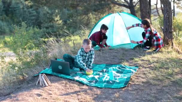 Lets go camping. Family camping. Reach destination place. Two girls pitch tent and one boy watching show on the laptop, sitting on the plaid. Wanderlust discovery. - Footage, Video