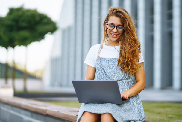 Smiling young woman in glasses with wireless headphones and a laptop working or studying online on the background of a city building. Concept for education, business, blog or freelance. - Foto, afbeelding