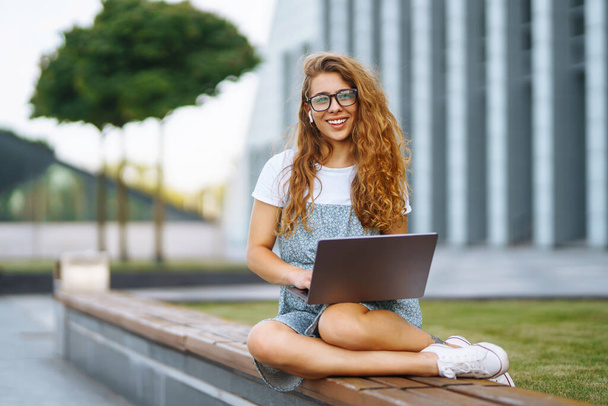 Smiling young woman in glasses with wireless headphones and a laptop working or studying online on the background of a city building. Concept for education, business, blog or freelance. - Zdjęcie, obraz