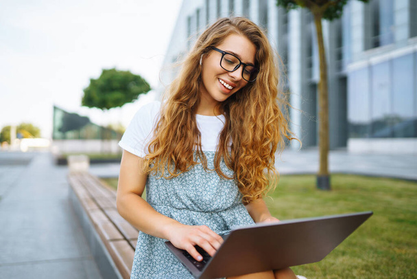 Smiling young woman in glasses with wireless headphones and a laptop working or studying online on the background of a city building. Concept for education, business, blog or freelance. - Photo, Image