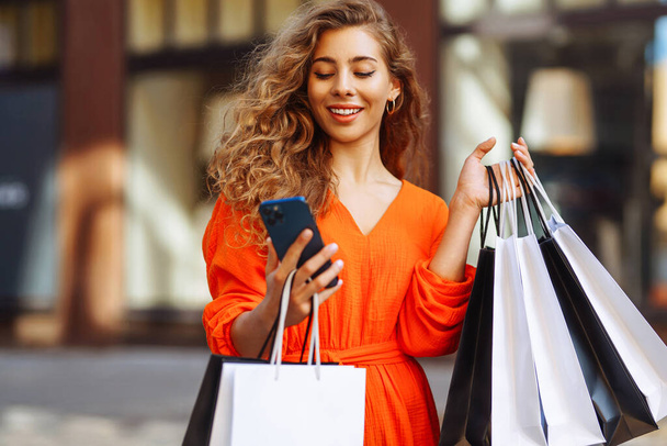 Happy female tourist doing shopping using smartphone while walking in sunny european city. Shopping lifestyle concept, consumerism. - Photo, image