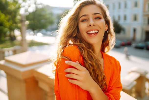 Beautiful young woman posing against backdrop of urban landscape in bright orange dress. Female tourist in bright dress enjoys sights of the city on sunny day. Concept of lifestyle, fashion, travel. - Photo, Image