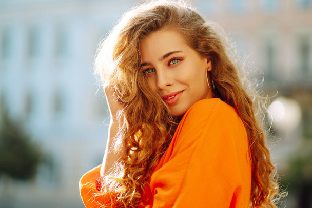 Beautiful young woman posing against backdrop of urban landscape in bright orange dress. Female tourist in bright dress enjoys sights of the city on sunny day. Concept of lifestyle, fashion, travel. - Fotó, kép