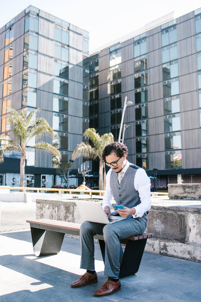 mladý latinský podnikatel pomocí notebooku a telefonu pracující na počítači v lavičce venku v Mexico City, Latinská Amerika hispánci - Fotografie, Obrázek