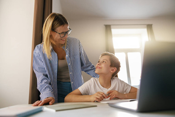 Sonriente mamá ayudando a su hijo haciendo la tarea de estudiar en línea utilizando el ordenador portátil en casa - Foto, Imagen