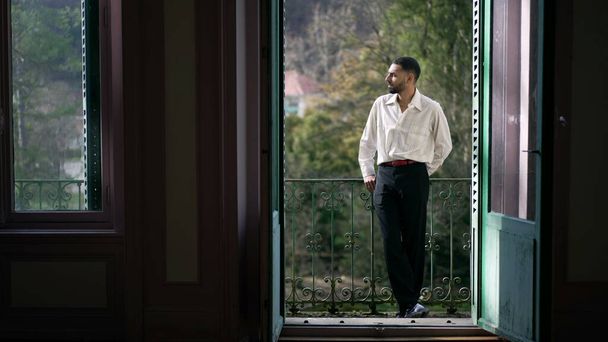 Meditasyon yapan genç bir adam evinin balkonunda dikilip temiz hava ve güneş ışığı alıyor. Düşünceli bir ifadeye sahip zarif bir Orta Doğulu erkek. - Fotoğraf, Görsel