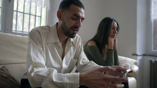 Ένα ζευγάρι σε κρίση. Αγνοεί την κοπέλα του ενώ κρατάει κινητό στο σπίτι. Φιλενάδα θέλει προσοχή από συνεργάτη απορροφάται από το περιεχόμενο των μέσων κοινωνικής δικτύωσης σε απευθείας σύνδεση κάθεται στον καναπέ σε εσωτερικούς χώρους - Φωτογραφία, εικόνα