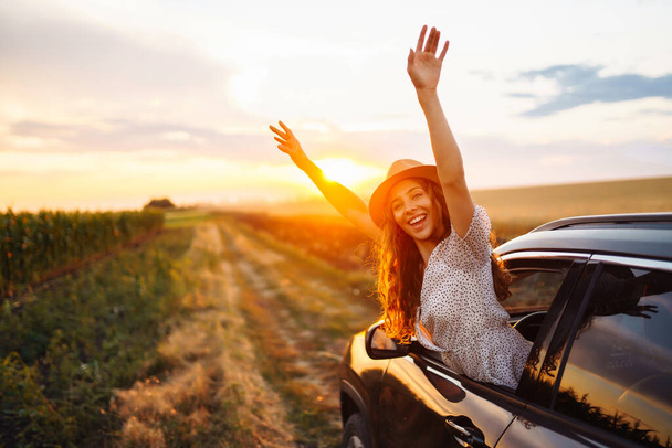 Young happy woman in hat enjoys car ride, leaning out of the window. In summer, a beautiful tourist woman travels by car and enjoys the golden sunset. Concept of an active lifestyle, travel, tourism. - 写真・画像