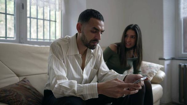 Couple in crisis. Man ignoring girlfriend while holding cellphone device at home. Girlfriend wants attention from partner absorbed by social media content online sitting on couch indoors - Photo, Image